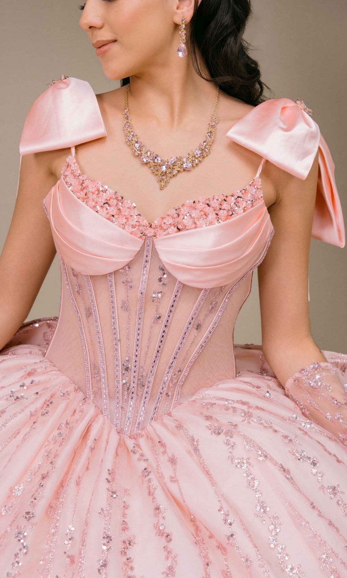 A woman wearing the Quinceanera Dress 8530JQ by Cinderella Couture, a pink sequined ball gown featuring satin bows on the shoulders and a beaded corset bodice, accessorized with a jeweled necklace and matching earrings, shown from the shoulders to the waist.